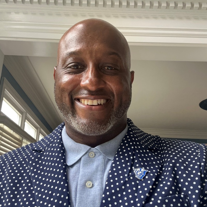 Smiling man wearing a blue polka dot blazer over a light blue shirt, standing indoors. The mood is cheerful and welcoming.