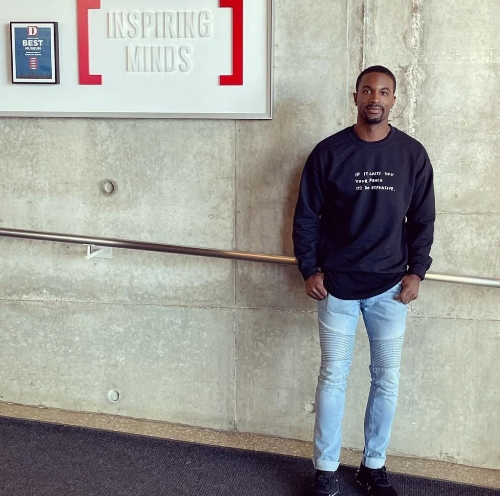 A man stands in front of a concrete wall and metal railing, wearing a black sweatshirt and light jeans. A sign nearby reads "Inspiring Minds." The mood is motivational.