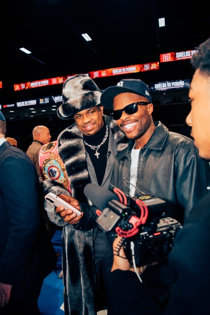 Two people posing smilingly for the camera; one in a luxurious fur coat and hat holding a championship belt, another wearing sunglasses and a leather jacket. A camera crew is in the foreground, capturing the moment in a lively, indoor setting.