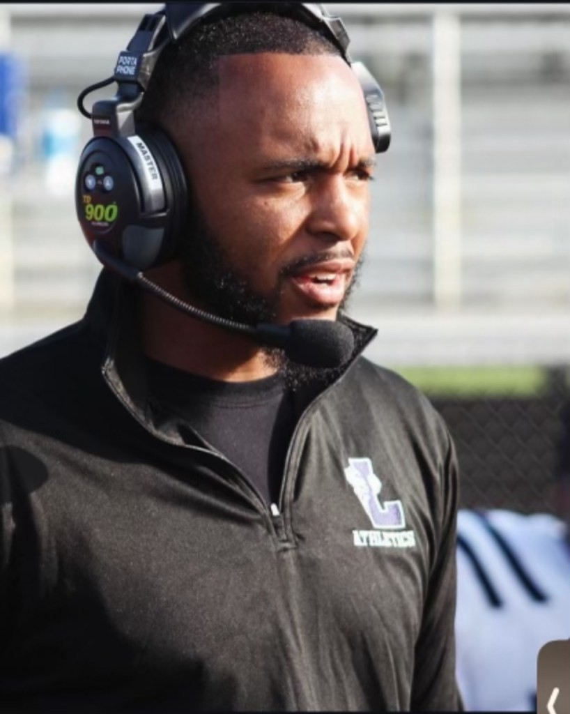 A man wearing a headset and dark athletic jacket stands on a sports field, focused and speaking, with blurred bleachers in the background.