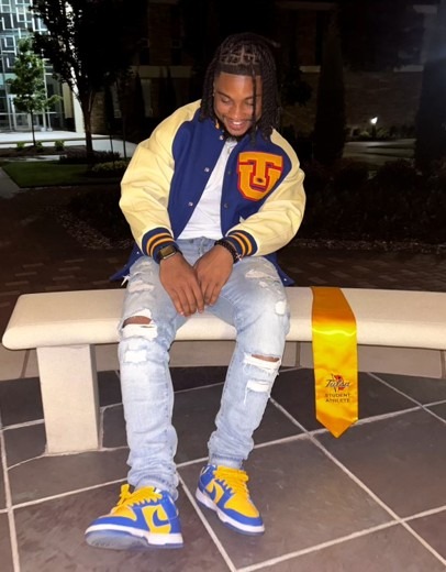 A person sits on a bench wearing a yellow and blue varsity jacket and shoes, with ripped jeans and a graduation stole. They are smiling, creating a joyful tone.