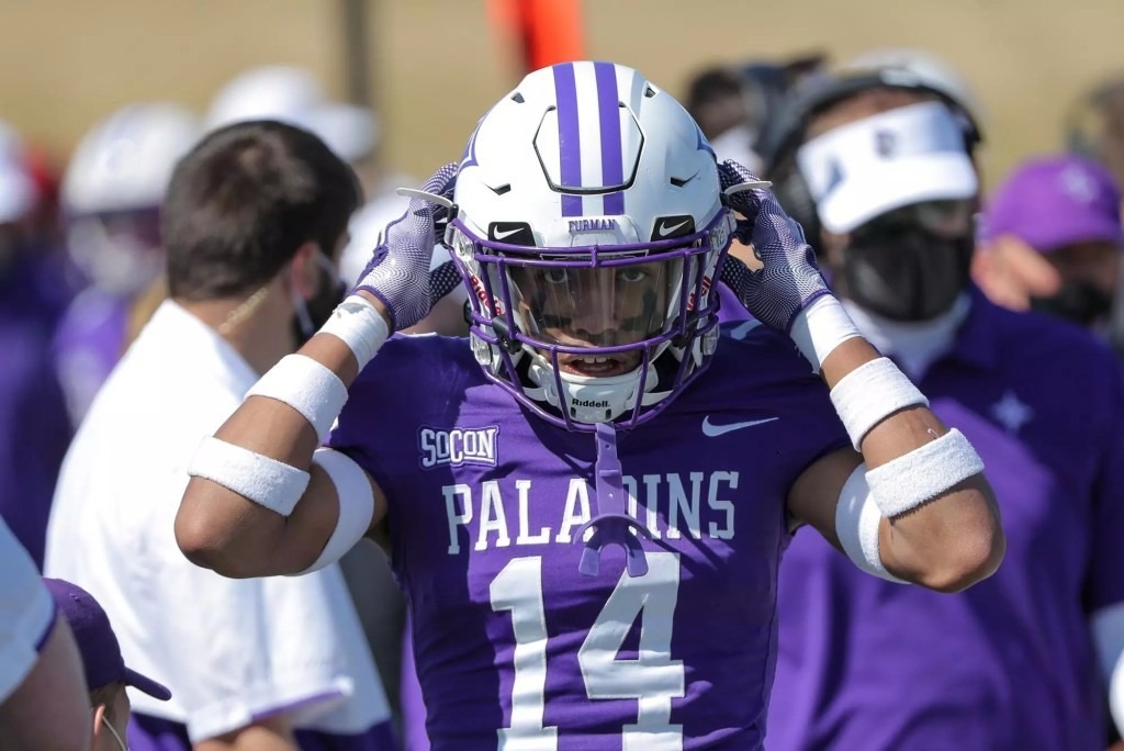 A football player in a purple jersey with "Paladins" and number 14 adjusts his helmet. The background shows blurred team staff wearing masks.