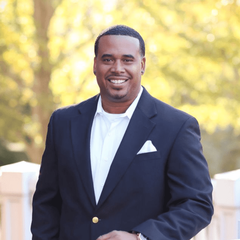 A smiling man in a black suit stands outdoors against a blurred backdrop of trees