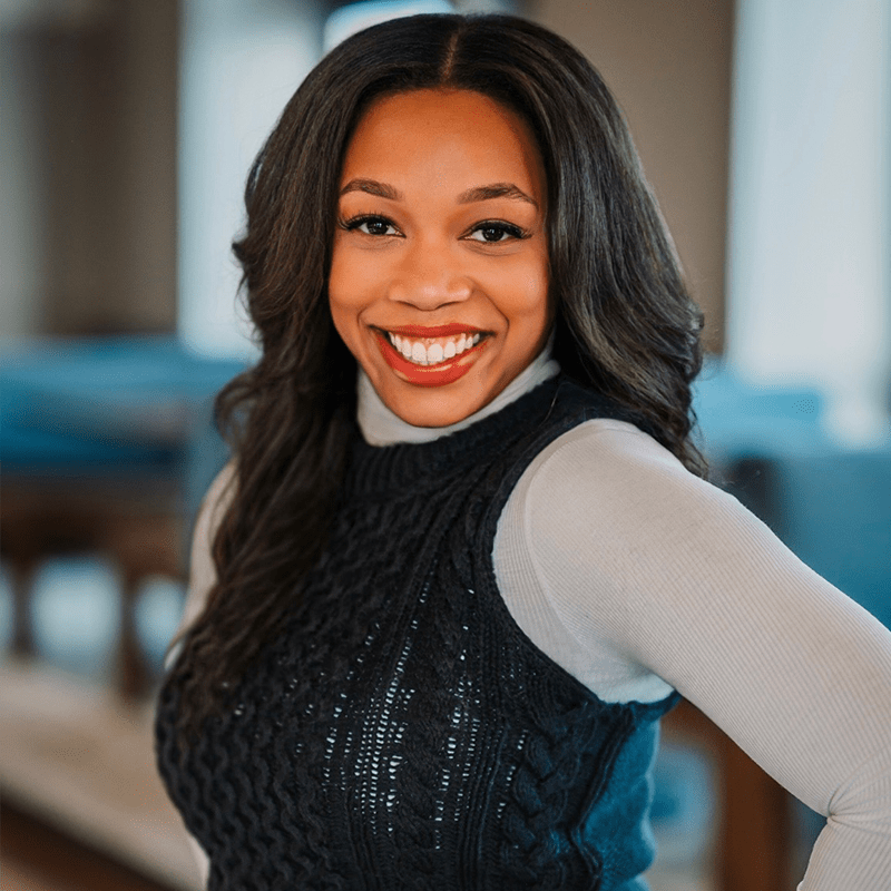 Young woman with long, wavy dark hair smiles brightly while wearing a knitted sleeveless top over a turtleneck