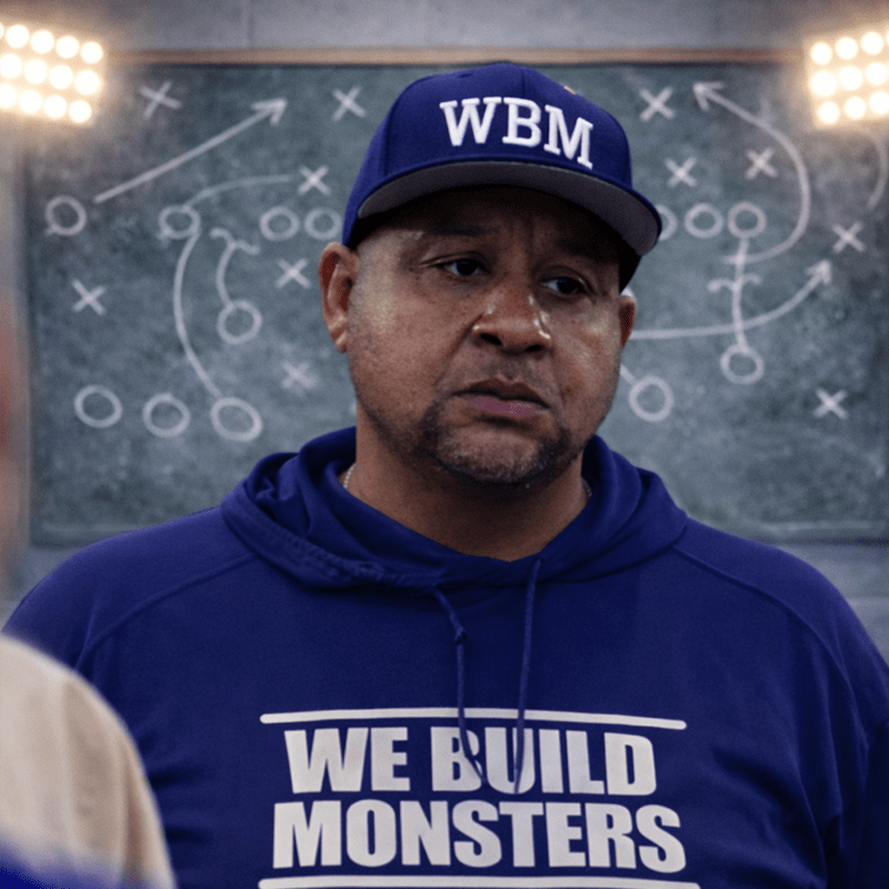 A focused coach wearing a blue hoodie with "WE BUILD MONSTERS" and a matching cap